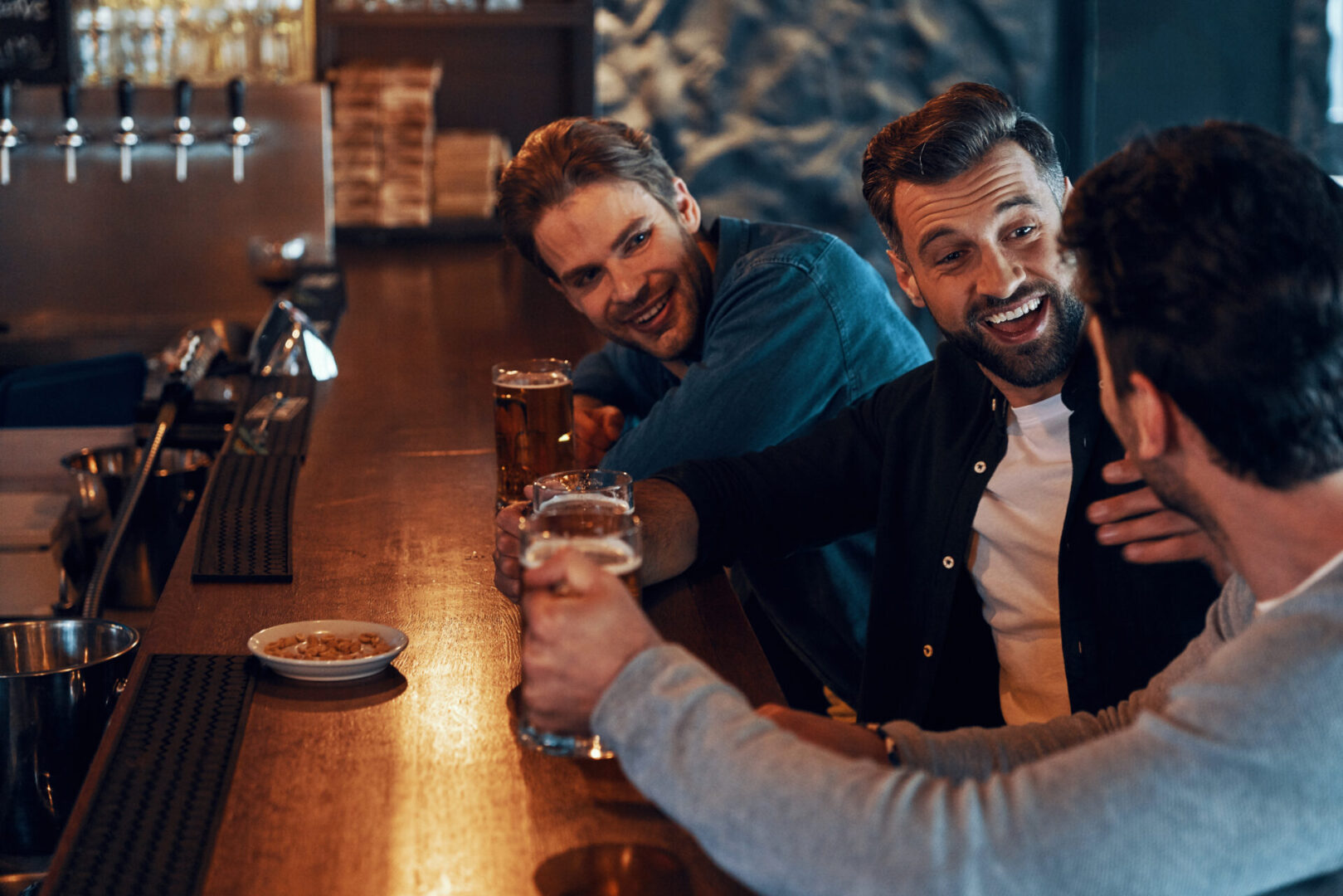 Carefree young men in casual clothing drinking beer and bonding together Three friends laughing at a bar counter.