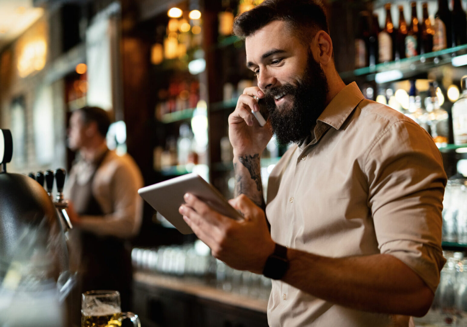 Young happy bartender using touchpad while talking on the phone. Man talking on phone, holding tablet.