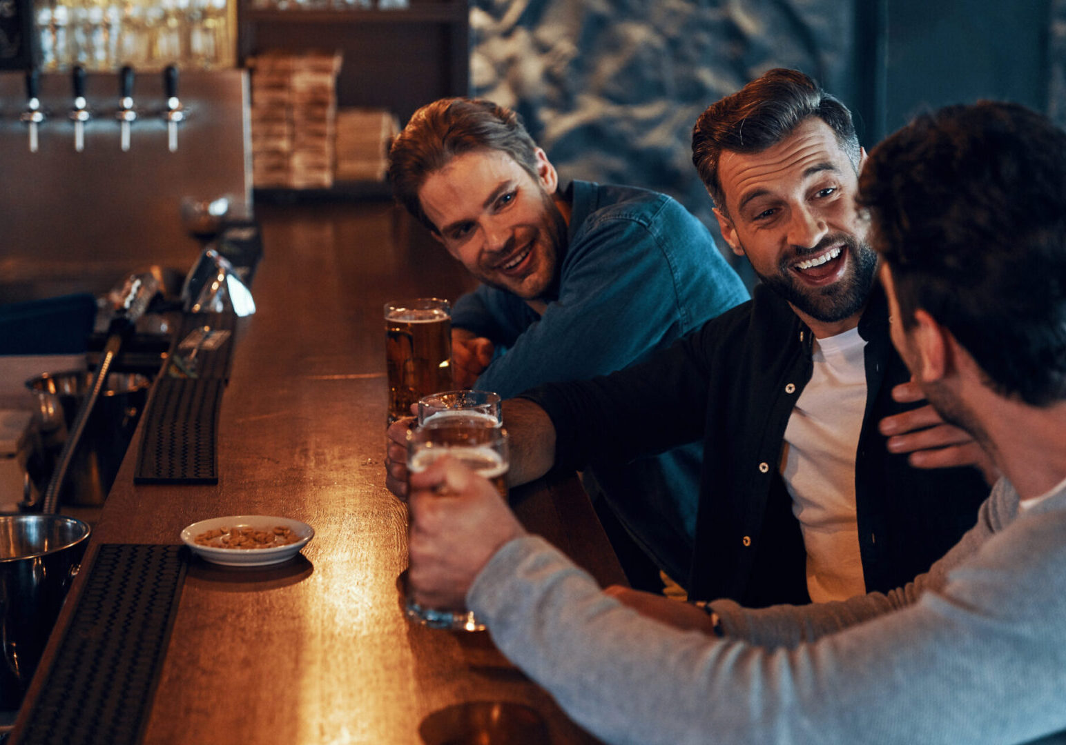 Carefree young men in casual clothing drinking beer and bonding together Three friends laughing at a bar counter.